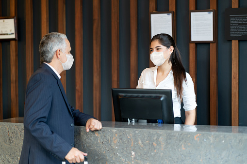 Friendly latin american hotel receptionist doing check in for male guest both wearing protective face masks - Coronavirus lifestyles