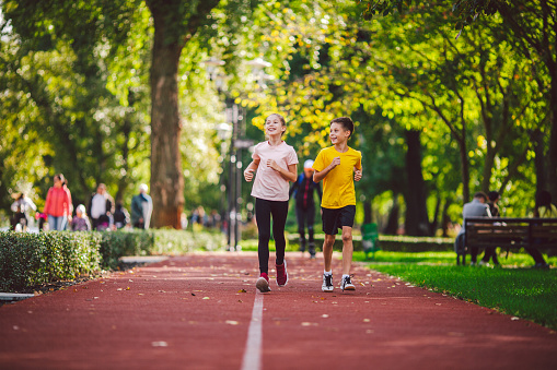 Couple of kids boy and girl doing cardio workout, jogging in park on jogging track red. Cute twins runing together. Run children, young athletes. Teen brother and sister running along path outdoors.