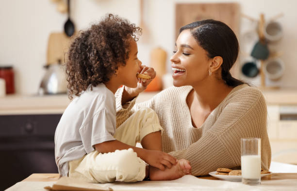 família afro-americana feliz: mãe e filho pequeno comem biscoitos com leite em casa - cookie women eating beautiful - fotografias e filmes do acervo