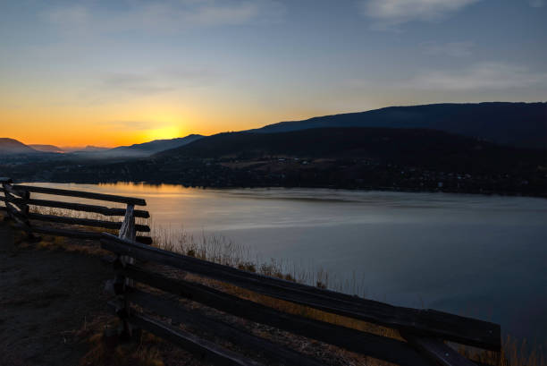 Wooden fence, fence in the foreground, water surface with a reflection of the sunrise, residential buildings located on the slope of the mountains, sun, and sky with clouds in the background.