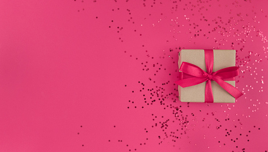 Gift box wrapped in craft paper with pink ribbon with bow and confetti on pink background. Monochrome festive flat lay with copy space.