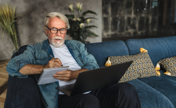 сосредоточенный пожилой человек, пишущий заметки в блокноте, наблюдая вебинар или онлайн обучение с помощью ноутбука современный старший � - computer old men laptop стоковые фото и изображения