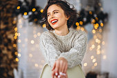 Pretty and happy brunette sitting in warm sweater on the chair with christmas lights behind.