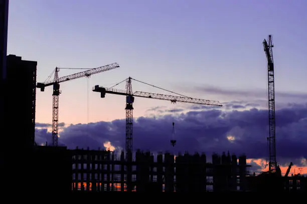 Night photography of a construction site, silhouettes of a lift-up in red and an unfinished residential building, gloomy dark blue sky sunset, construction industry