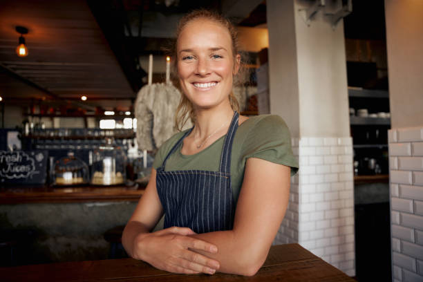 istock Cheerful young female waitress wearing apron standing leaning on table in cafe 1282265365