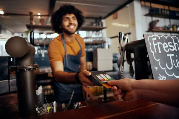 Photo of Waiter holding credit card swipe machine while customer typing code in modern cafe