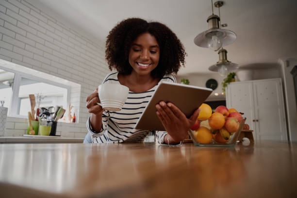 istock Smiling african woman standing in kitchen leaning on counter using digital tablet while drinking coffee 1282271234
