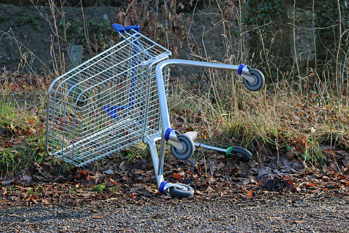 Abandoned metal supermarket trolley with blue plastic handle, lying on the side at the edge of a road with a grass verge and trees in the background.
