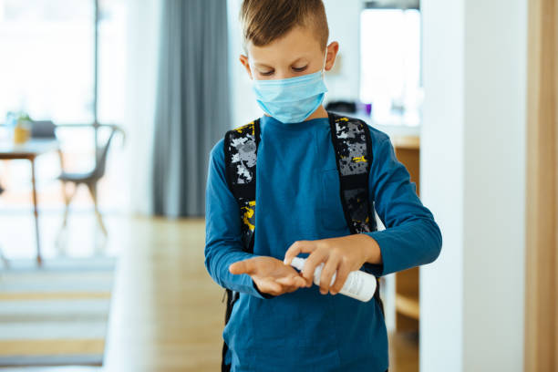 Schoolboy with protective face mask spraying hand sanitizer to his hands indoors Schoolboy with protective face mask spraying hand sanitizer to his hands indoors backpack sprayer stock pictures, royalty-free photos & images