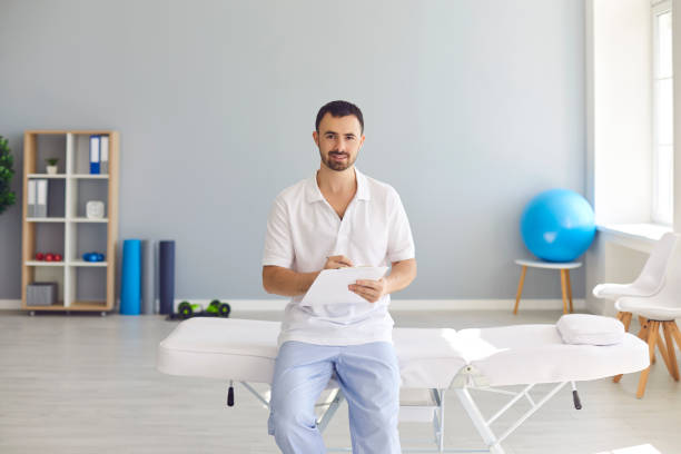 jeune docteur mâle avec le presse-papiers s’asseyant sur le lit d’hôpital dans le bureau de la nouvelle clinique - massagist photos et images de collection