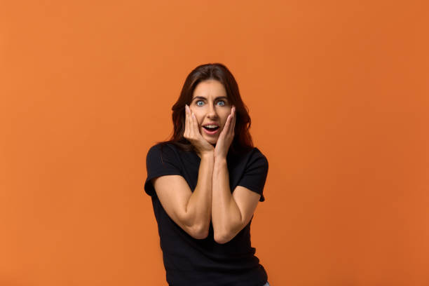 istock Joyful amazed female keeps mouth opened, hands on cheeks, expresses great surprisment, being shocked by saw something, stares at camera. Isolated over an orange background. 1282283267