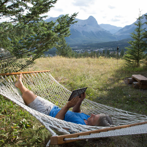 Man relaxes in hammock and uses digital tablet in mountain meadow Mountains in the distance hammock men lying down digital tablet stock pictures, royalty-free photos & images