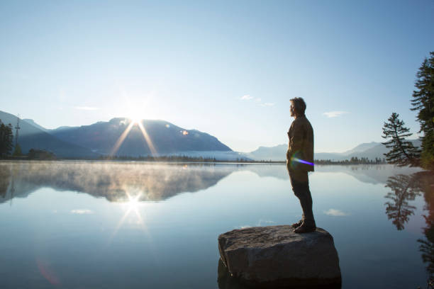 hombre maduro se para en la roca, en el lago, y mira el amanecer - dawn mountain range mountain canadian rockies fotografías e imágenes de stock