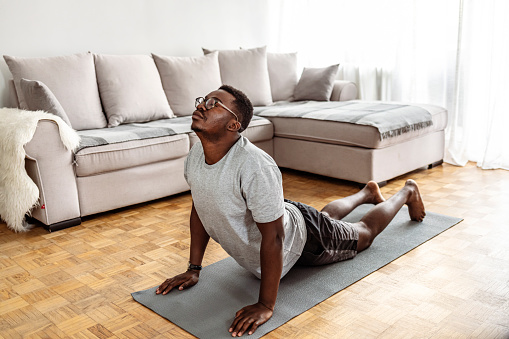 Young man in cobra yoga pose on exercise mat doing stretching exercise in his living room during Covid-19 pandemic lockdown.
