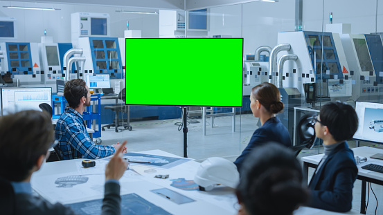 Modern Factory Office Room: Diverse Team of Engineers, Managers and Investors Sitting at Meeting Table, They wave at Interactive TV that shows Green Screen Video Call Conference Template