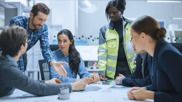 istock Busy Diverse Team of Computer Engineers, Specialists Gather Around Conference Table, They Discuss Project Drafts, Blueprints, Find Problem Solutions. Electronic Technology Factory Office Meeting Room 1282300414