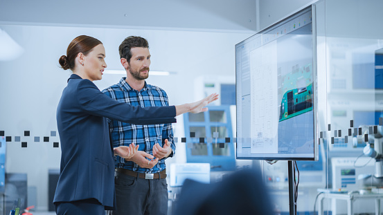 Female Computer Engineer and Male Project Manager Use Digital Interactive Whiteboard That Shows 3D Printed Circuit Board Concept. Developers Find Problem Solution. Modern Factory Meeting Room