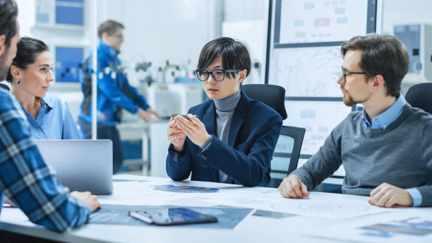 istock Factory Office Meeting Room: Computer Design Specialist Holds Printed Circuit Board Prototype and Reports to Diverse Team of Engineers, Managers Sitting at Conference Table. Industrial Facility 1282300437
