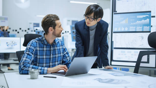 istock Modern Factory: Industrial Engineer Sitting at His Desk, Working on Laptop Computer, Talks with Chief Project Manager, They Analyze Mechanisms and Blueprints. Manufactory with CNC Working Machinery 1282300444