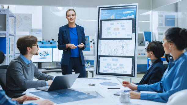 istock Modern Industrial Factory Meeting: Confident Female Engineer Uses Interactive Whiteboard, Makes Report to a Group of Engineers, Managers Talks and Shows Concept of Sustainable Eco-Friendly Engine 1282300486
