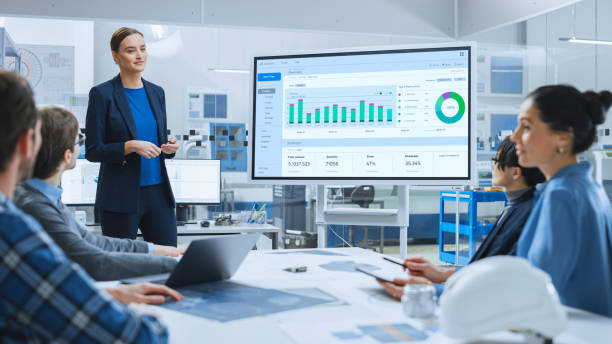 istock Modern Industrial Factory Meeting: Confident Female Engineer Uses Interactive Whiteboard, Makes Report to a Group of Engineers, Managers Talks and Shows Statistics, Growth and Analysis Information 1282301032