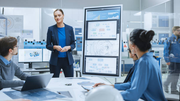 istock Modern Industrial Factory Meeting: Confident Female Engineer Uses Interactive Whiteboard, Makes Report to a Group of Engineers, Managers Talks and Shows Concept of Sustainable Eco-Friendly Engine 1282301033