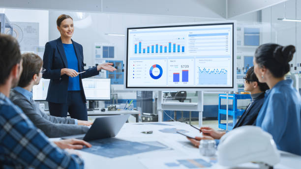istock Modern Industrial Factory Meeting: Confident Female Engineer Uses Interactive Whiteboard, Makes Report to a Group of Engineers, Managers Talks and Shows Statistics, Growth and Analysis Information 1282301087