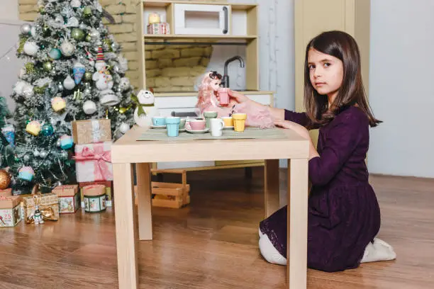 Seven-year-old beautiful dark-haired girl plays with doll at tea party table in Christmas room