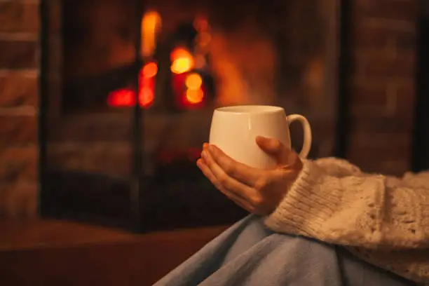Young woman wearing white woolen knitted sweater enjoying hot tea near fireplace in a cozy living room in the evening.