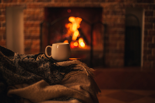 Mug with hot tea standing on a chair with woolen blanket in a cozy living room with fireplace.