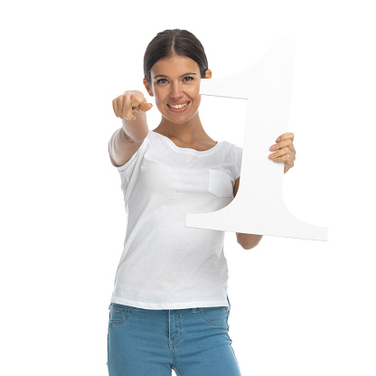 Positive casual woman holding number one and pointing while standing on white studio background