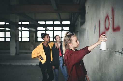 Group of teenagers girl gang standing indoors in abandoned building, using spray paint on wall.