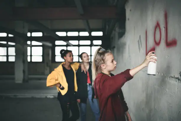 Photo of Group of teenagers girl gang indoors in abandoned building, using spray paint on wall.