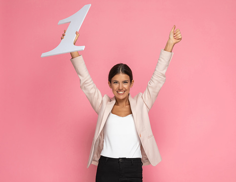beautiful businesswoman holding a number one and giving a thumbs up in the air against pink background