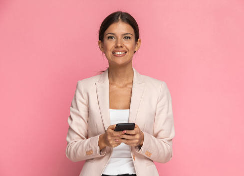 beautiful businesswoman texting on her phone and smiling at the camera on pink background