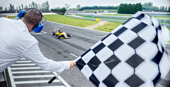 Rear view of auto race official waving chequered flag to yellow open-wheel single-seater racing car cars driving on track.