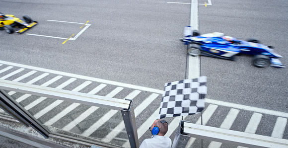 High angle view of auto race official waving chequered flag to blue and yellow open-wheel single-seater racing car cars driving on track.