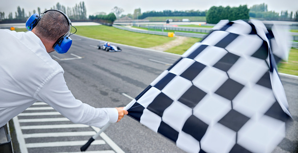 Rear view of auto race official waving chequered flag to blue open-wheel single-seater racing car car driving on track.