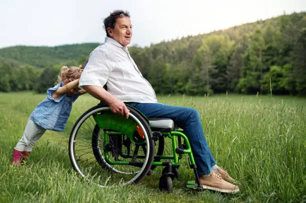 Photo of Small girl with senior grandfather in wheelchair on a walk on meadow in nature.