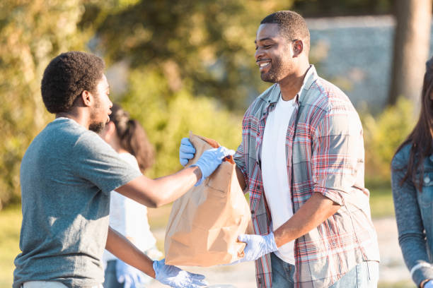 Food bank volunteer hands bag to young man in need A cheerful mid adult male food bank volunteer hands a paper bag filled with donated food items to a young man. The young man has taken a financial loss during the COVID-19 pandemic. giving tuesday stock pictures, royalty-free photos & images