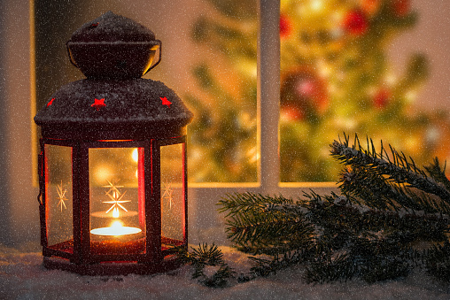 Red lantern on an outside widowsill covered in snow on a snowing night. A Christmas tree inside the house in visible through the window. Copy space.