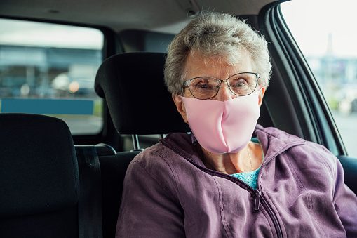 A medium, front view of a senior, Caucasian woman sat waiting in the car for their carer to return. She is wearing a face mask to protect themselves and others form Covid-19.