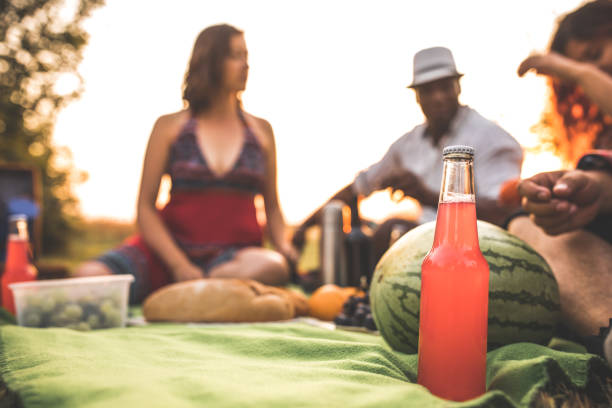 Focus on foreground of colorful refreshing drink in glass bottle placed next to a watermelon. Multicultural friends are having a picnic.