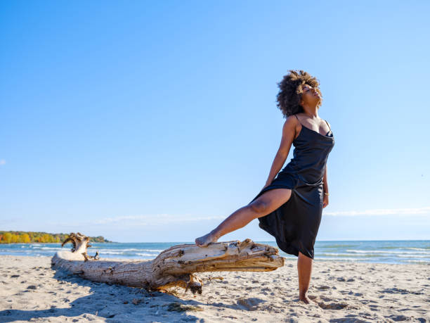 Young African woman wearing black dress, relaxing on the sandy beach. Sunny day in October.