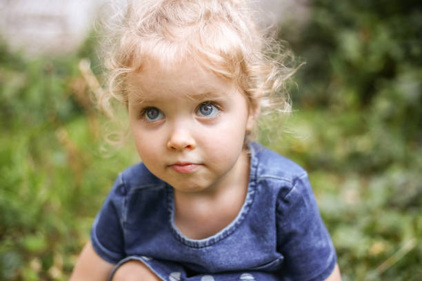 verticale d’une petite belle fille européenne avec les cheveux blonds bouclés et les yeux bleus de 3 à 4 ans dans une robe bleue classique de couleur, couleur tendance 2020. la fille joue dans le jardin dehors au printemps ou en été - child caucasian little girls 3 4 years photos et images de collection