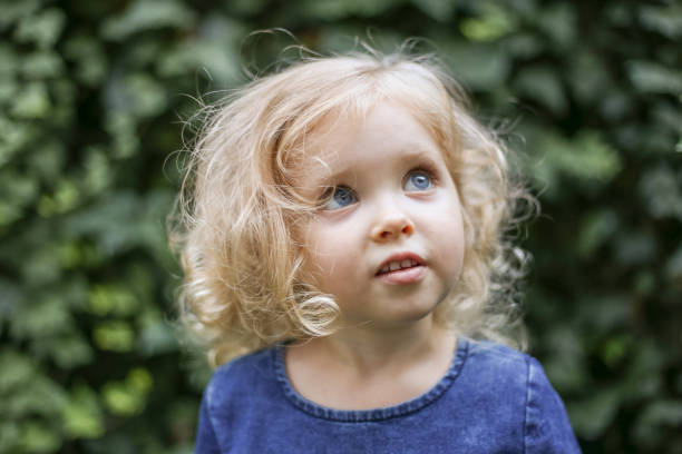 portrait of a little beautiful european girl with curly blonde hair and blue eyes of 3 to 4 years in a classic blue color dress, trendy color 2020. girl plays in the garden outside in spring or summer - child caucasian little girls 3 4 years imagens e fotografias de stock