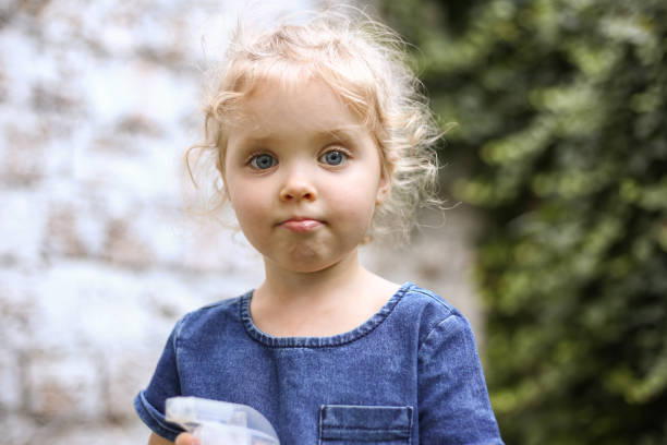 verticale d’une petite belle fille européenne avec les cheveux blonds bouclés et les yeux bleus de 3 à 4 ans dans une robe bleue classique de couleur, couleur tendance 2020. la fille joue dans le jardin dehors au printemps ou en été - child caucasian little girls 3 4 years photos et images de collection