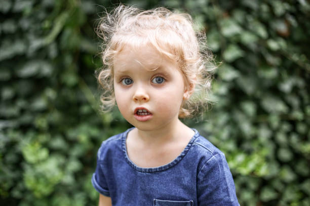 verticale d’une petite belle fille européenne avec les cheveux blonds bouclés et les yeux bleus de 3 à 4 ans dans une robe bleue classique de couleur, couleur tendance 2020. la fille joue dans le jardin dehors au printemps ou en été - child caucasian little girls 3 4 years photos et images de collection