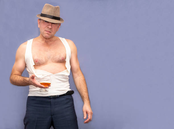 帽子と破れたtシャツを着た男が、ウイスキーやコニャックのグラスを持って壁に立っています。 - eastern european caucasian one person alcoholism ストックフォトと画像