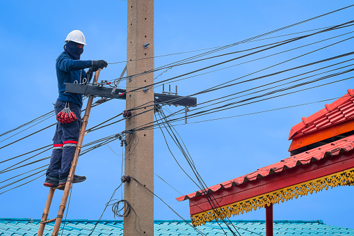 Asian technician on wooden ladder is installing cable lines to connect telephone and internet signal system on electric pole against blue sky background